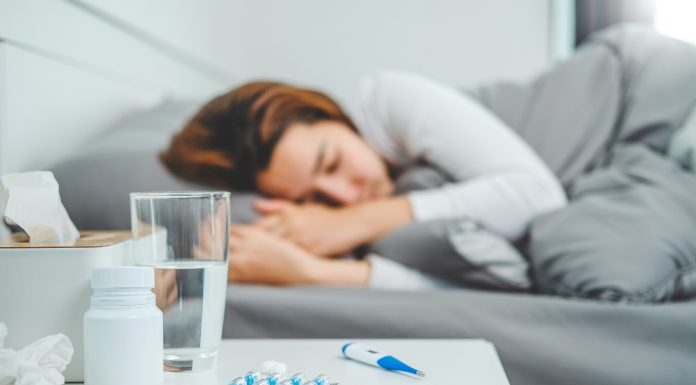 Oseba z gripo leži v postelji z vročino, ob njej zdravila, termometer in kozarec vode